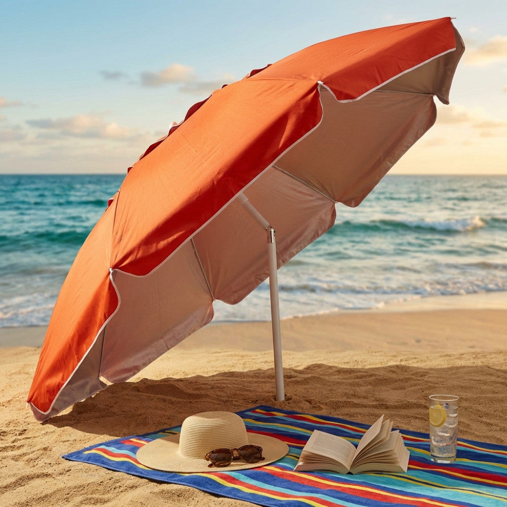 Ombrellone da spiaggia arancione con dettagli bianchi aperto sulla sabbia, affiancato da un telo mare a righe, un cappello di paglia, occhiali da sole e un libro aperto.