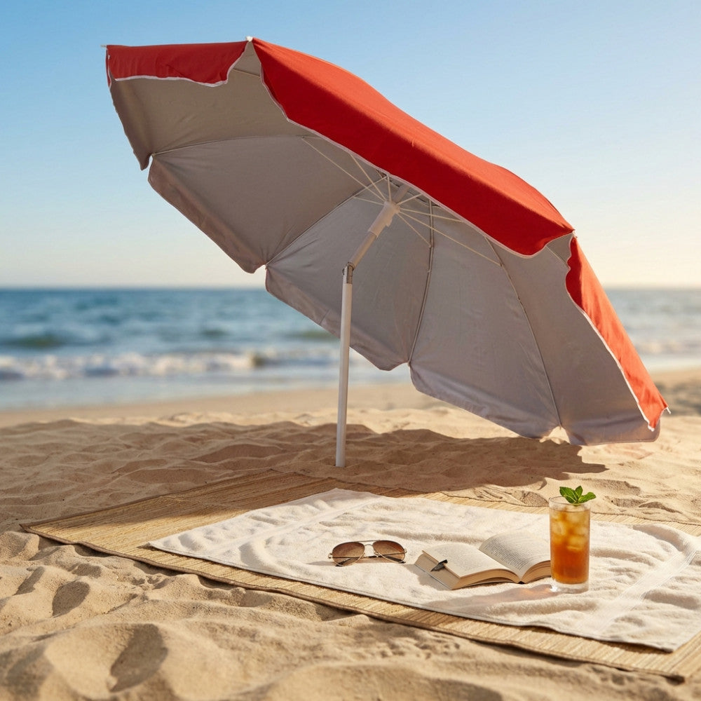 Ombrellone da spiaggia arancione inclinato sulla sabbia, che proietta ombra su una stuoia intrecciata con un asciugamano bianco, un libro e un cocktail ghiacciato.