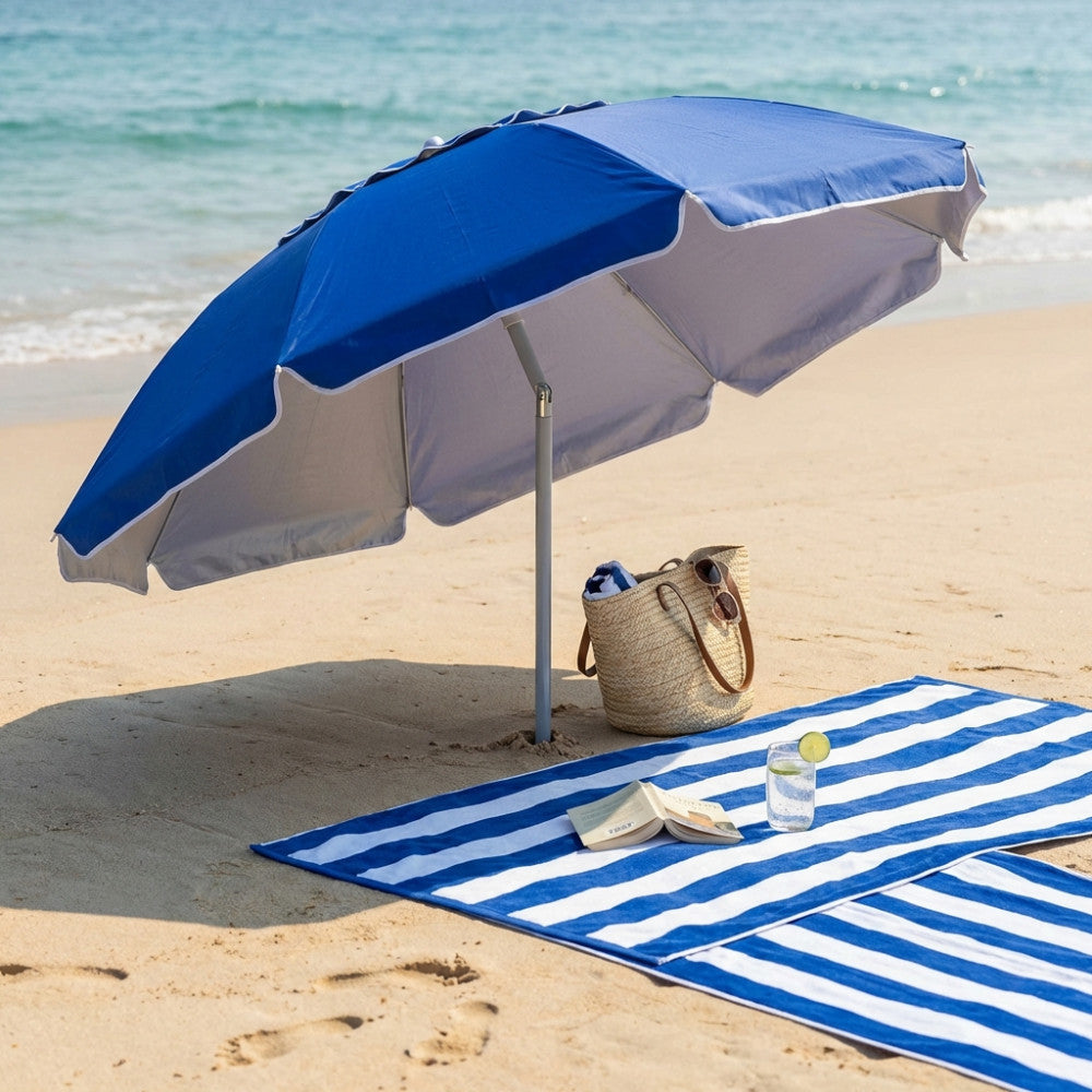 Ombrellone da spiaggia blu OM807041 inclinato sulla sabbia, con telo mare a righe bianche e blu, libro e borsa in paglia.
