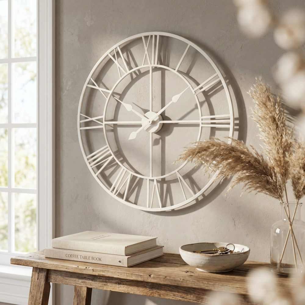 Grande orologio da parete a scheletro in metallo bianco con numeri romani, appeso sopra una consolle in legno rustico decorata con libri e pampas.