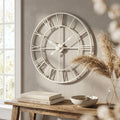 Grande orologio da parete a scheletro in metallo bianco con numeri romani, appeso sopra una consolle in legno rustico decorata con libri e pampas.