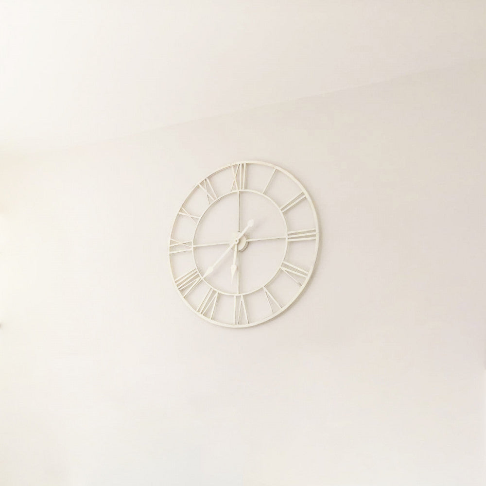 Orologio da parete in metallo bianco dal design minimalista e vintage, appeso in alto su una parete bianca e pulita.