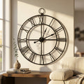Orologio da parete M Constant in metallo antracite con numeri romani, appeso sopra una consolle in legno illuminata dalla luce naturale.