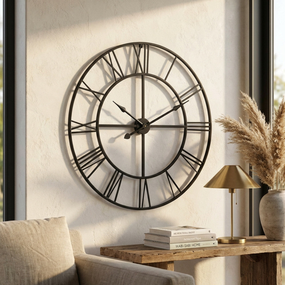 Orologio da parete in metallo antracite con numeri romani, appeso sopra una consolle in legno grezzo con lampada in ottone.
