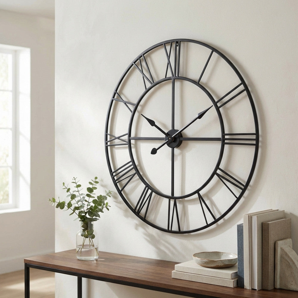 Orologio S Constant in metallo scheletrato su parete bianca, abbinato a un vaso in vetro con rami di eucalipto.