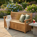 Panca contenitore Surat in teak posizionata in un giardino fiorito con cuscini verdi e una coperta chiara.