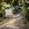 Panca Dollie in acciaio bianco ossidato ambientata in un giardino fiorito con cuscino e libro.
