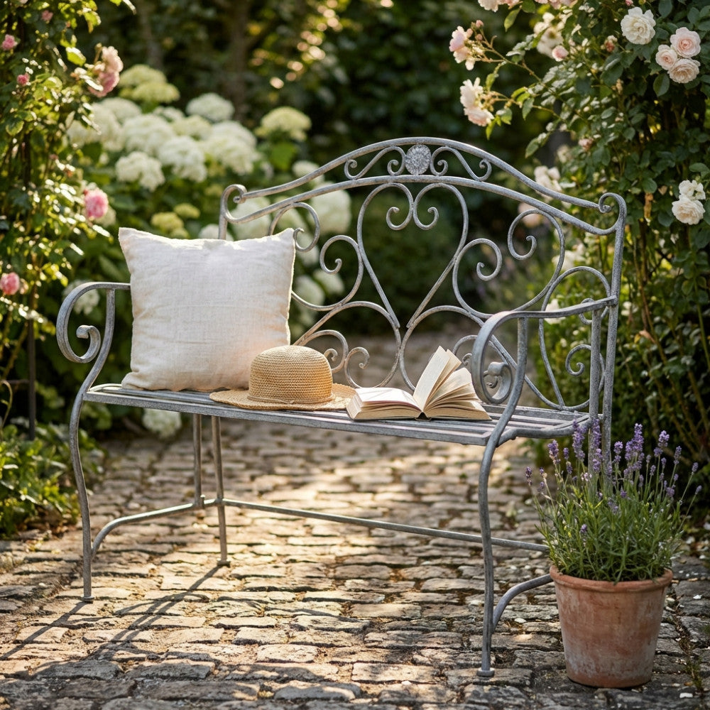 Panca Dollie in acciaio bianco ossidato ambientata in un giardino fiorito con cuscino e libro.