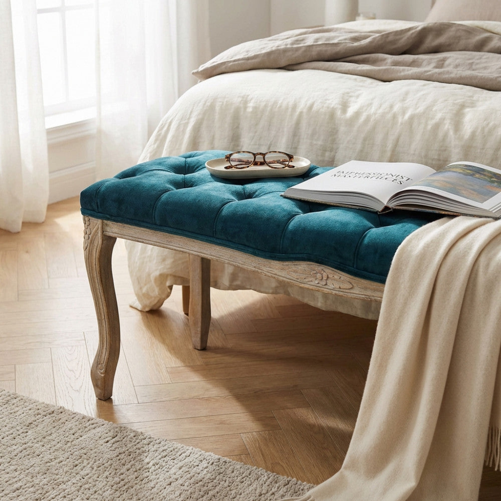 Panca Misty in velluto blu posizionata ai piedi di un letto con lenzuola in lino, decorata con un libro e occhiali.