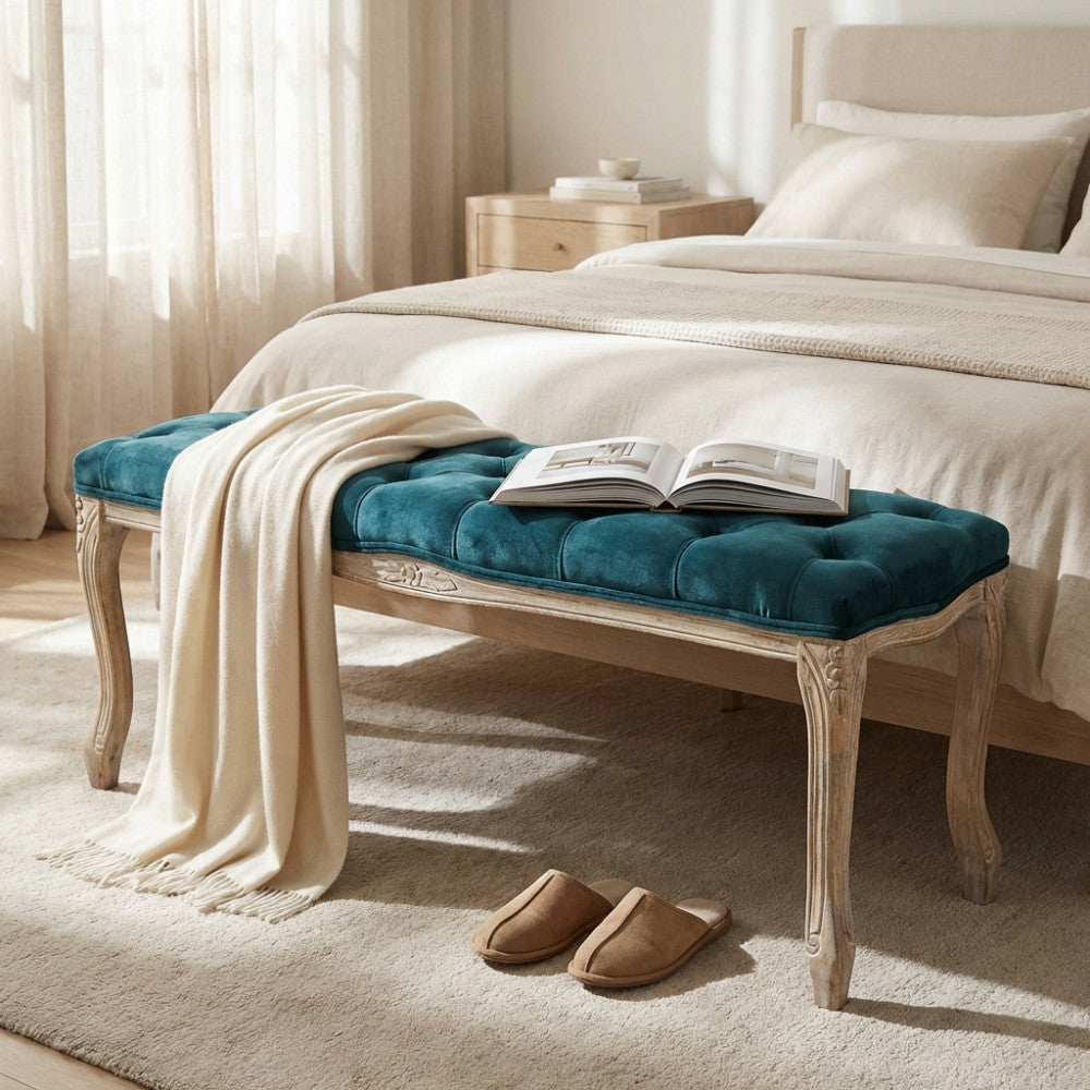 Vista angolare della panca in velluto blu e legno intagliato in una camera da letto luminosa, con una coperta chiara appoggiata.