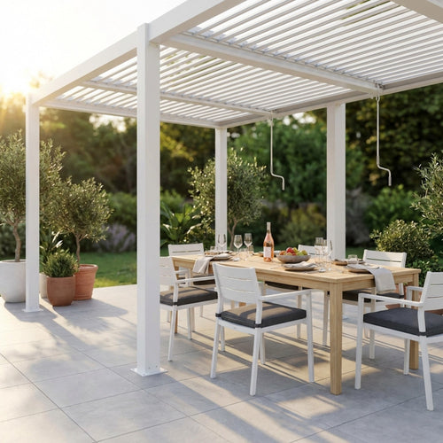 Pergola bioclimatica in alluminio bianco allestita con tavolo da pranzo in legno e sedie bianche in un giardino soleggiato.