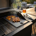 Piastra in ghisa per barbecue con spiedini di carne e verdure in cottura, fumo aromatico e birra ghiacciata sullo sfondo.