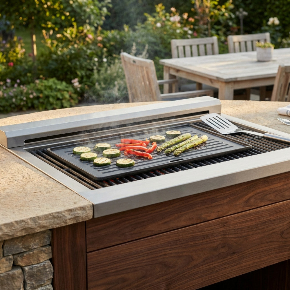 Dettaglio di una piastra in ghisa premium integrata in una cucina da esterno in pietra e legno, circondata da un giardino lussureggiante durante il tramonto.