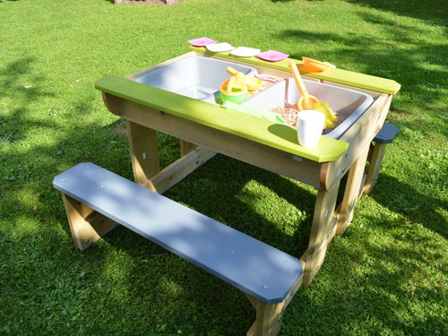 Tavolo da picnic per bambini in pino con piano aperto, che rivela due vasche integrate contenenti sabbia, acqua e giochi colorati, posizionato su un prato verde.