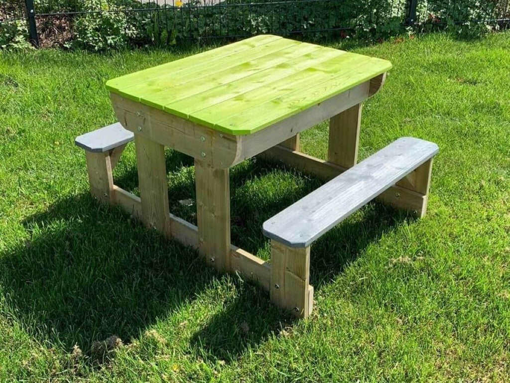 Tavolo da gioco per bambini con struttura in legno naturale, sedute grigie e piano chiuso color verde mela, immerso in un giardino soleggiato.