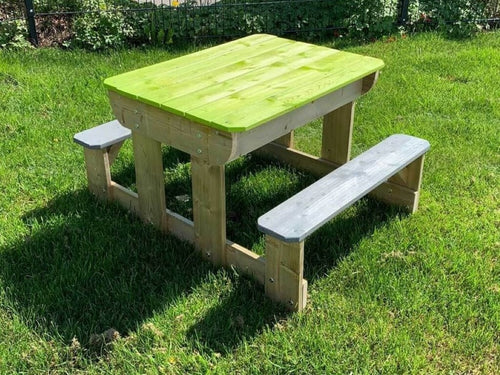 Tavolo da gioco per bambini con struttura in legno naturale, sedute grigie e piano chiuso color verde mela, immerso in un giardino soleggiato.
