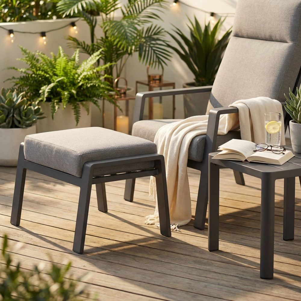 Poggiapiedi Giacarta in alluminio antracite e tessuto grigio, posizionato su un patio in legno accanto a una poltrona lounge.
