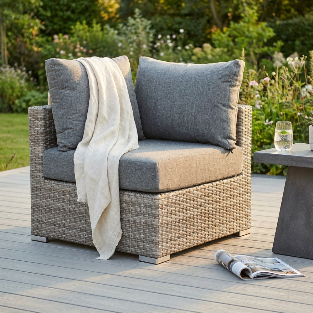 Poltrona angolare Copacabana in wicker grigio con cuscini e un morbido plaid, ambientata su un patio in legno al tramonto.