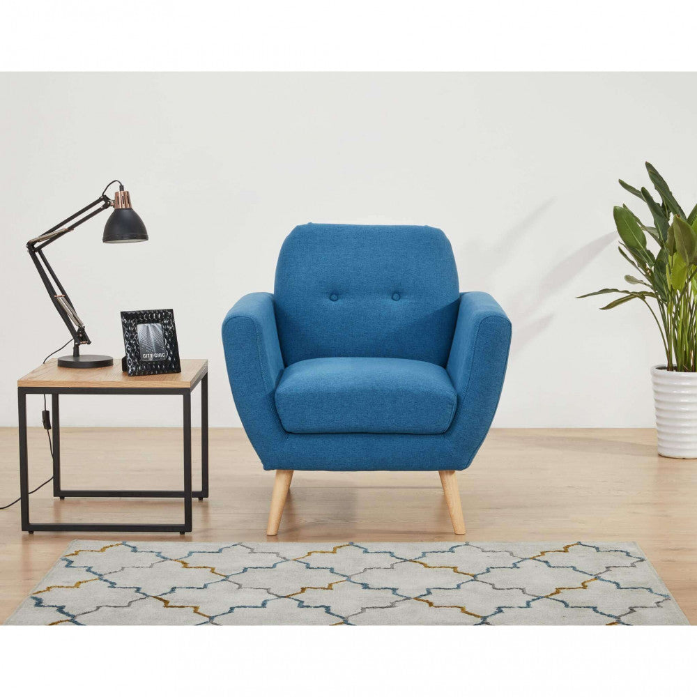 Vista frontale della poltrona Burton in tessuto blu inserita in un ambiente domestico moderno, con tappeto a motivi geometrici e lampada da tavolo.