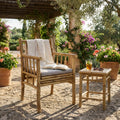 Poltrona CARLIS in bambù naturale con cuscino grigio chiaro, allestita in un soleggiato giardino mediterraneo con tavolino abbinato.