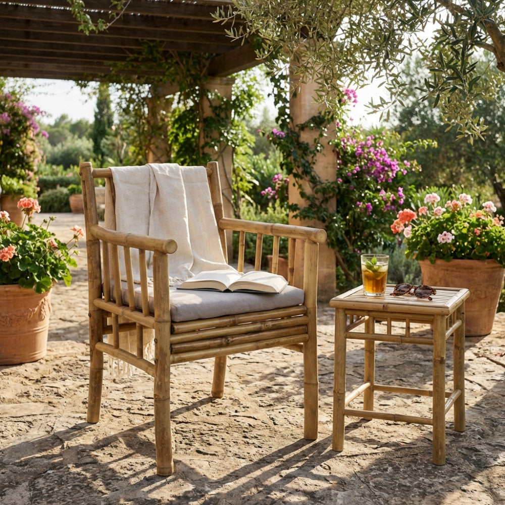 Poltrona CARLIS in bambù naturale con cuscino grigio chiaro, allestita in un soleggiato giardino mediterraneo con tavolino abbinato.