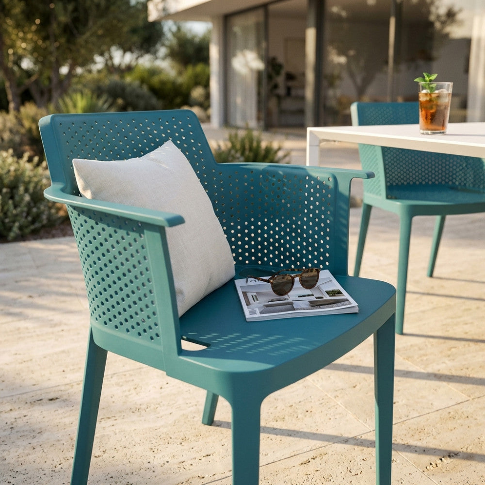 Poltrona outdoor Cathy verde petrolio con cuscino decorativo bianco, riviste e occhiali da sole su un patio.