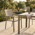 Poltrona Cervia in alluminio e polywood taupe ambientata in un giardino mediterraneo accanto a un tavolo.