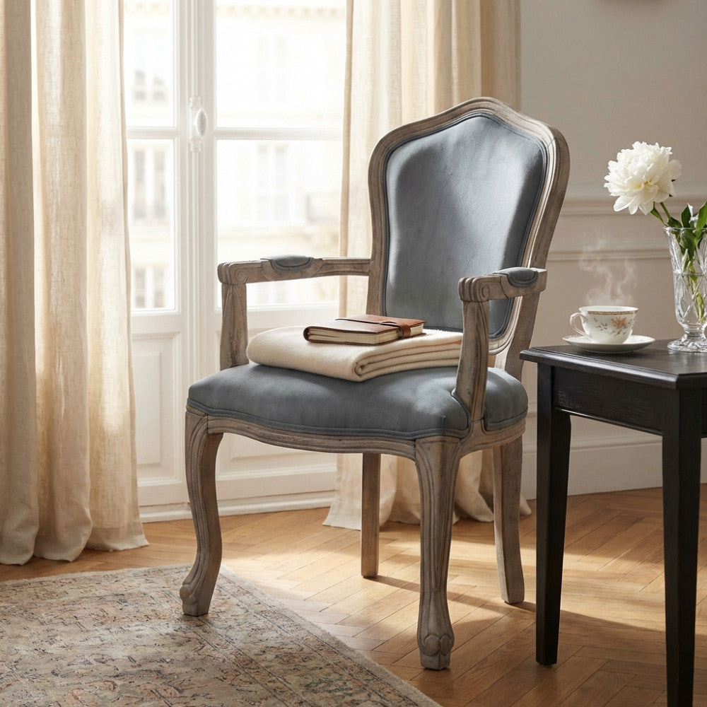 Poltrona classica in velluto grigio posizionata vicino a una finestra luminosa, con una coperta piegata e un tavolino nero laterale.