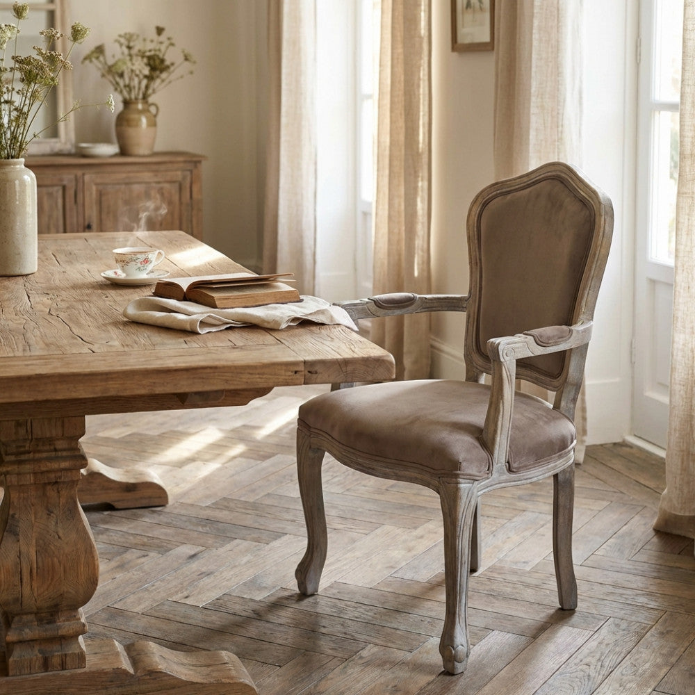 Poltrona Charlene in velluto taupe e legno intagliato, ambientata in una sala da pranzo rustica chic accanto a un tavolo in legno massello.