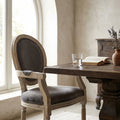 Poltrona Constance in velluto taupe e legno, posizionata accanto a un tavolo da pranzo rustico con un bicchiere d'acqua e un libro aperto.