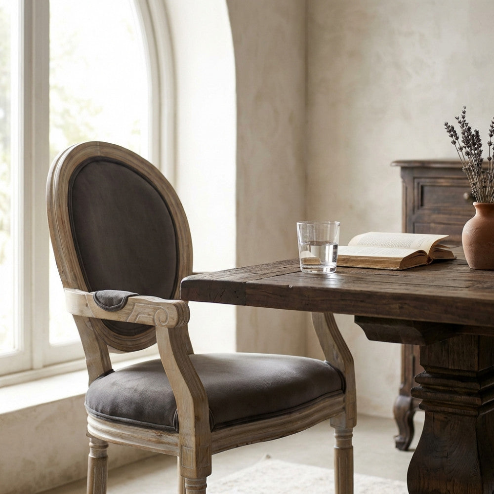 Poltrona Constance in velluto taupe e legno, posizionata accanto a un tavolo da pranzo rustico con un bicchiere d'acqua e un libro aperto.