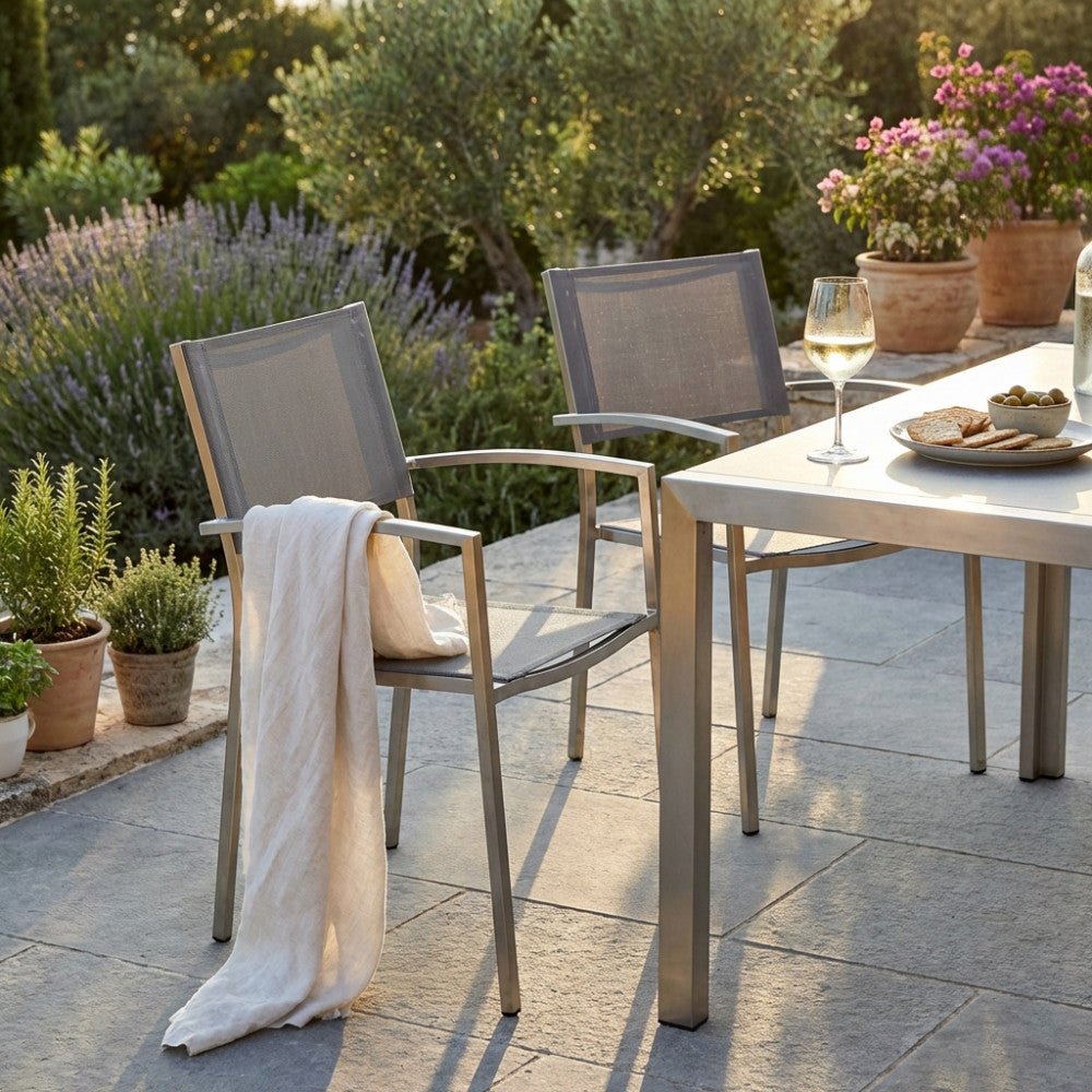 Poltrona Florida in acciaio e textilene grigio ambientata in un giardino lussuoso al tramonto, affiancata a un tavolo da pranzo outdoor.