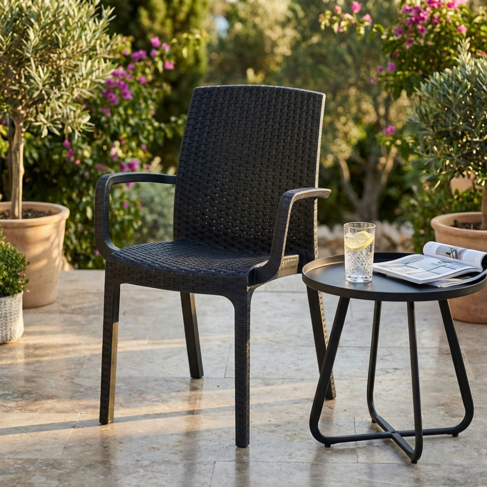 Poltrona Indiana in wicker antracite ambientata in un patio esterno soleggiato, affiancata da un tavolino nero con bicchiere d'acqua.