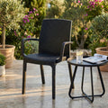 Poltrona Indiana in wicker antracite ambientata in un patio esterno soleggiato, affiancata da un tavolino nero con bicchiere d'acqua.