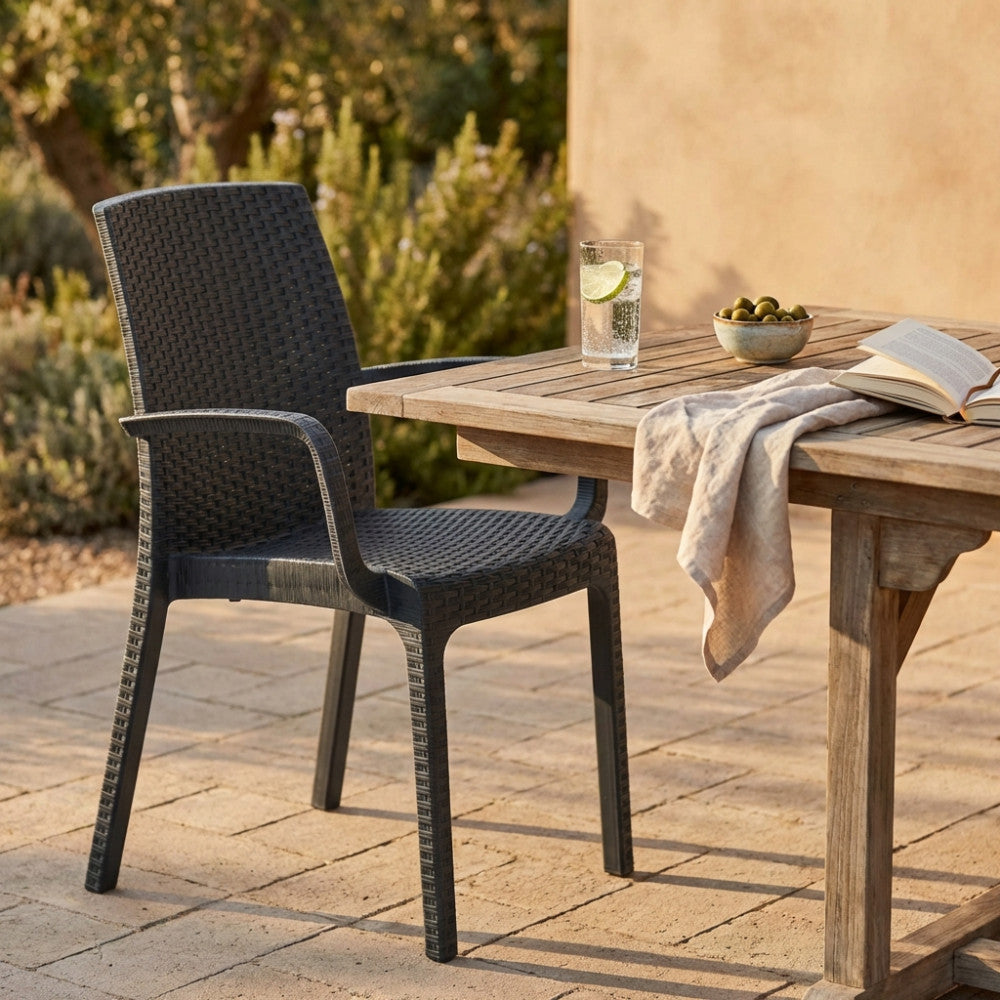 Poltrona con braccioli in wicker antracite posizionata accanto a un tavolo da pranzo in legno massello in un giardino mediterraneo.