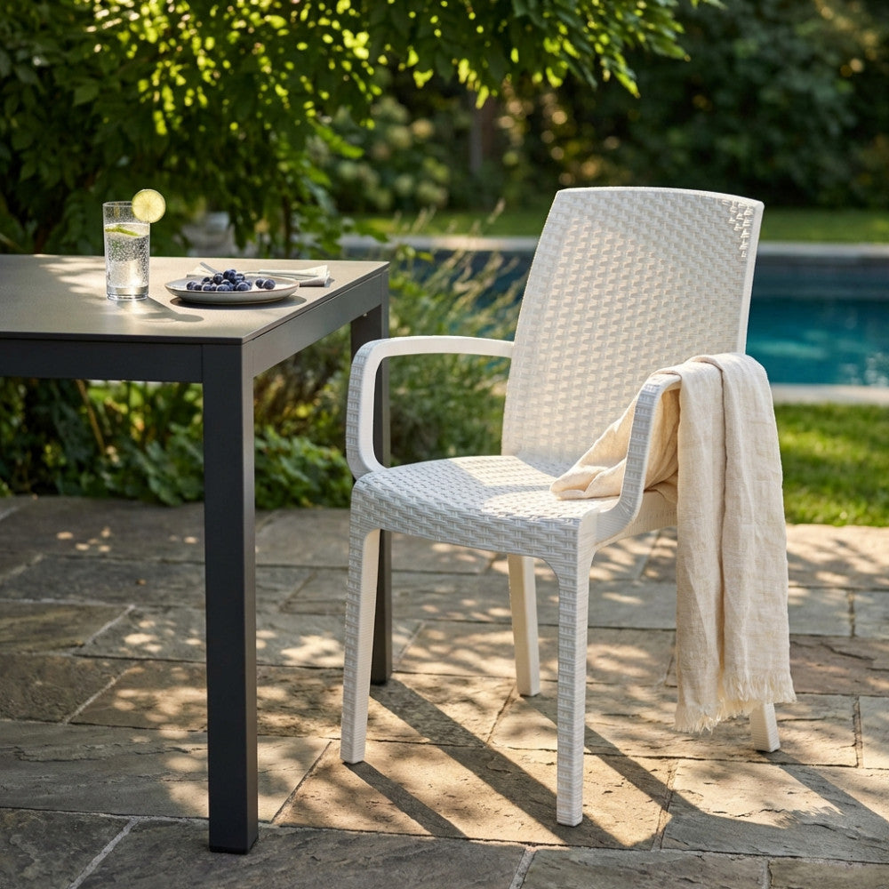 Poltrona Indiana in wicker bianco con braccioli, ambientata a bordo piscina vicino a un tavolo scuro.