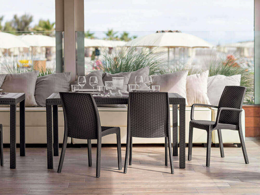 Allestimento outdoor di lusso con diverse poltrone Indiana in wicker marrone attorno a tavoli scuri sotto un porticato.