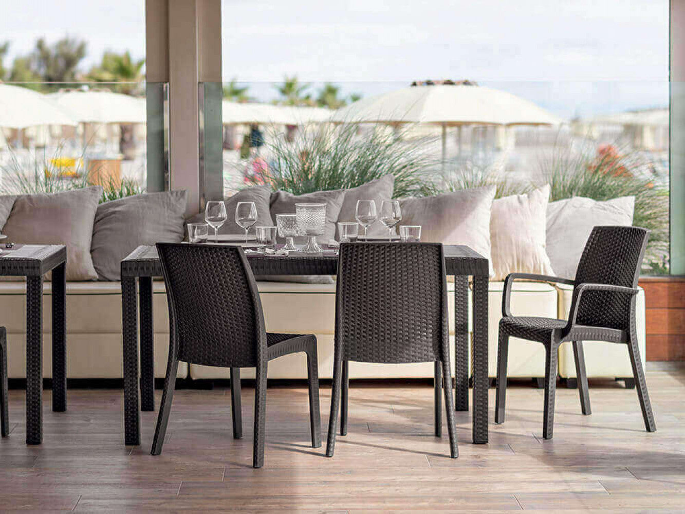 Allestimento outdoor di lusso con diverse poltrone Indiana in wicker marrone attorno a tavoli scuri sotto un porticato.
