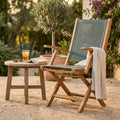 Poltrona Lahore pieghevole in teak e textilene grigio, ambientata in un rigoglioso giardino accanto a un tavolino in legno.