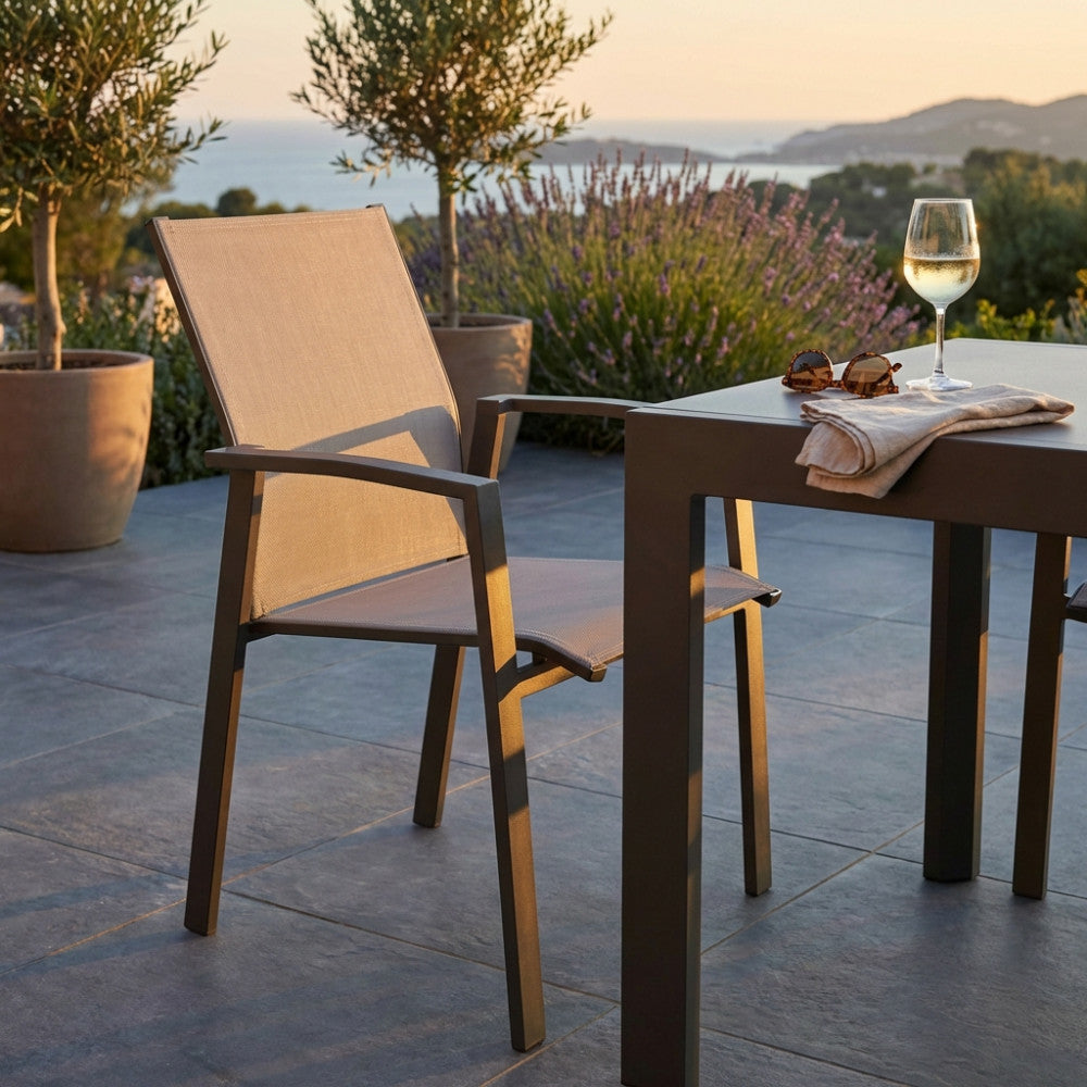 Poltrona Maili in alluminio e textilene color taupe ambientata su una terrazza esterna al tramonto, affiancata a un tavolo coordinato.