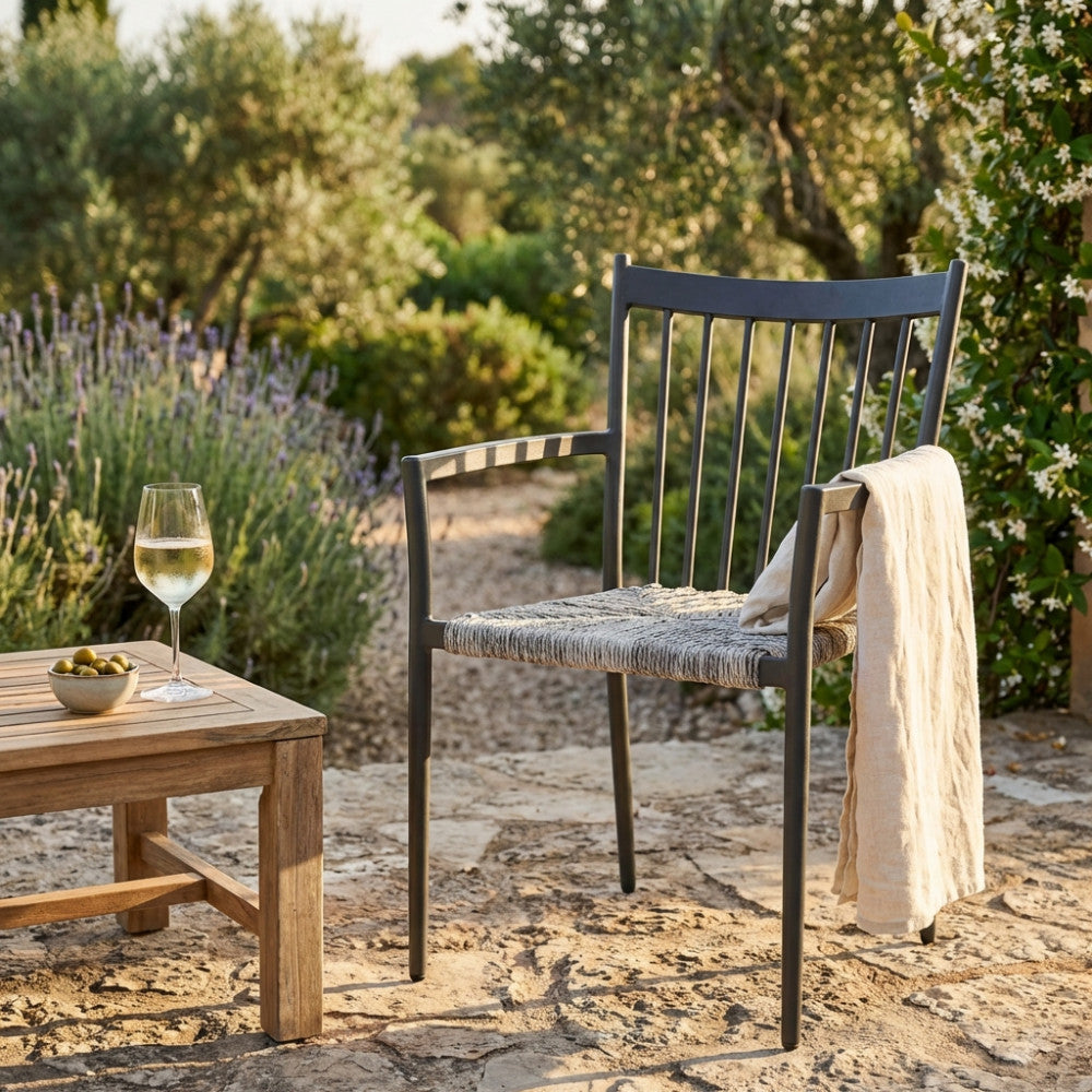 Poltrona Martinica in alluminio e wicker antracite ambientata in un giardino rustico con tavolino in legno e calice di vino.