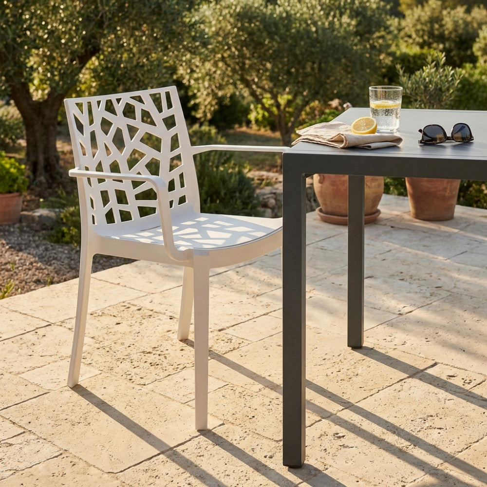 Poltrona Matrix bianca con braccioli posizionata accanto a un tavolo da pranzo in metallo scuro in un giardino mediterraneo.
