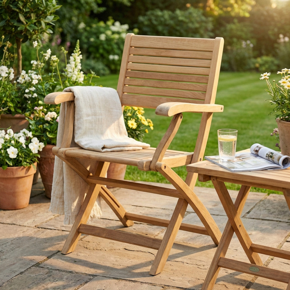 Poltrona Mirage in legno teak pieghevole, ambientata in un rigoglioso giardino con un telo in lino appoggiato sul bracciolo.