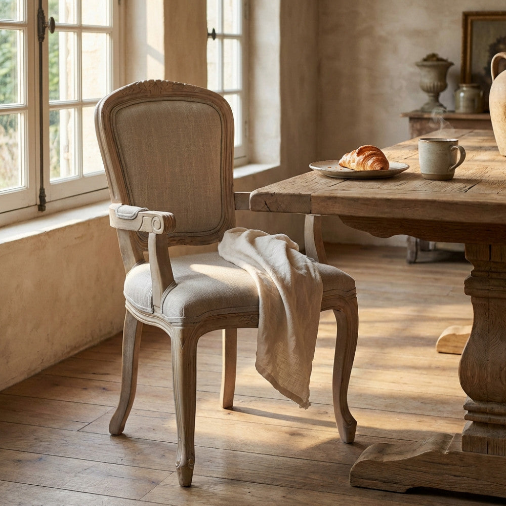 Poltrona Romy in tessuto grigio e legno intagliato, ambientata in una sala da pranzo rustica vicino a un tavolo in legno.
