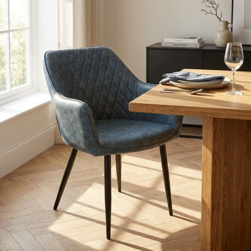 Poltrona Sindy in ecopelle blu posizionata accanto a un tavolo da pranzo in legno di rovere, illuminata dalla luce naturale di una finestra.