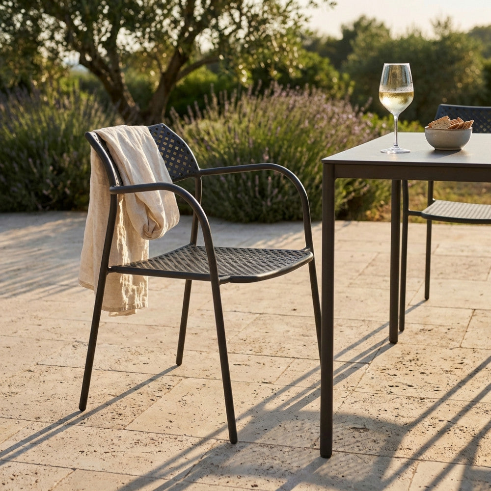 Poltrona Windsor in acciaio taupe ambientata in un patio esterno soleggiato, affiancata a un tavolo coordinato con un calice di vino e un telo in lino appoggiato sul bracciolo.