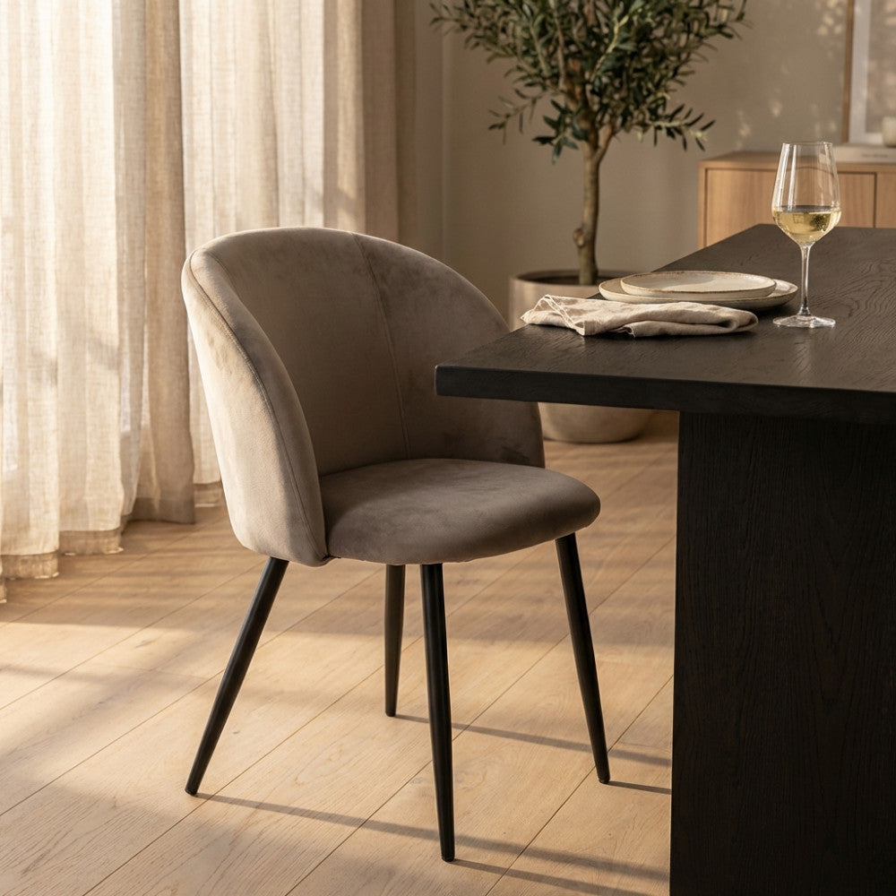 Elegante poltrona moderna in velluto tortora posizionata in una sala da pranzo raffinata, con calice di vino sul tavolo e ulivo decorativo sullo sfondo.