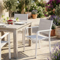 Poltrona Zante in alluminio bianco e textilene grigio ambientata in un rigoglioso giardino soleggiato accanto a un tavolo in legno.
