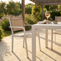 Poltrona Zante in alluminio e textilene color tortora, ambientata all'aperto accanto a un tavolo bianco con un calice di vino.