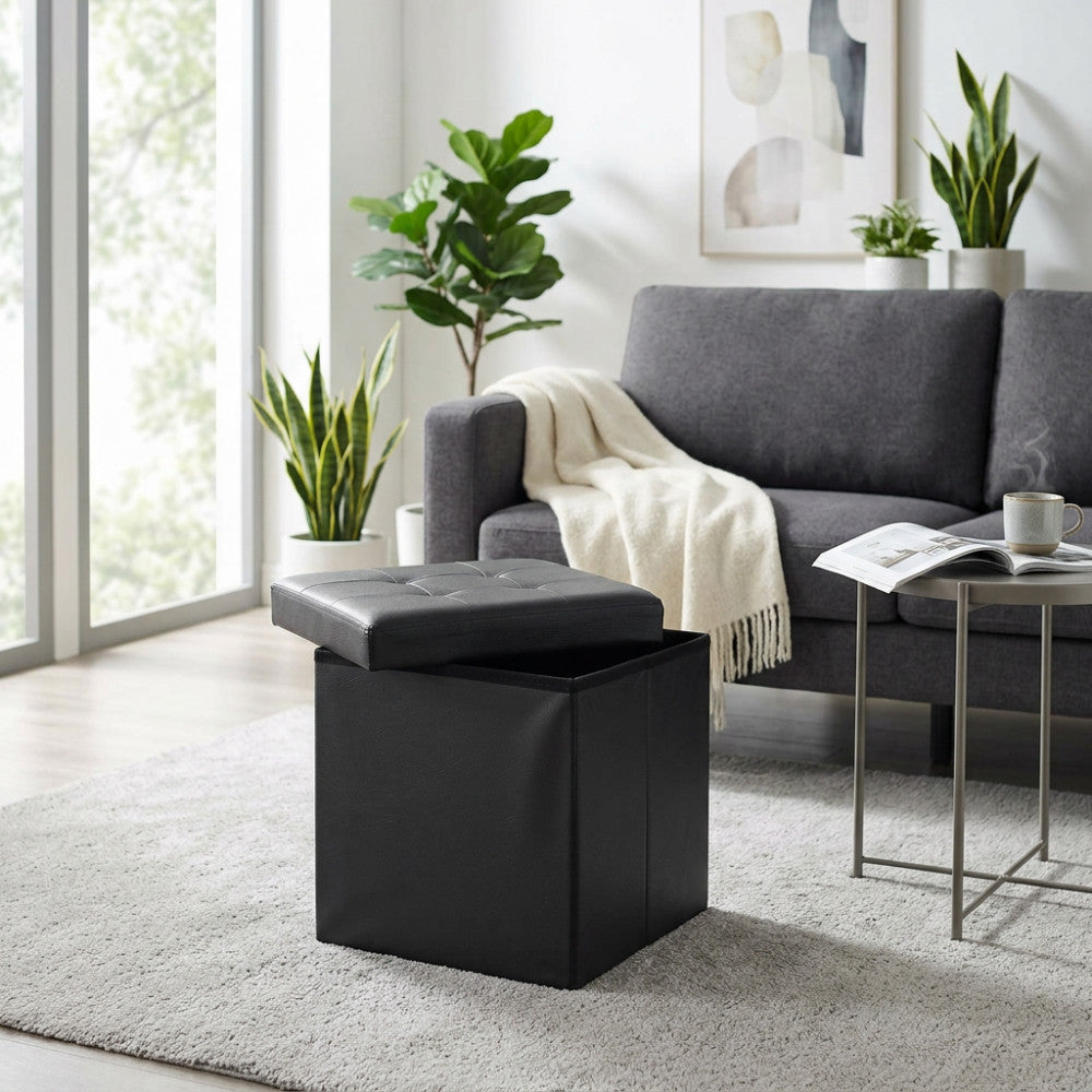 Pouf contenitore quadrato in ecopelle nera con coperchio semiaperto, posizionato su un tappeto chiaro accanto a un divano grigio in un salotto luminoso.